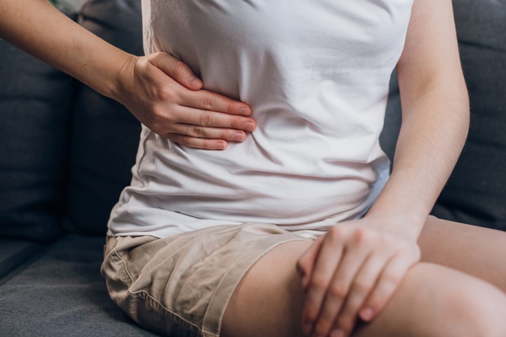 Close up of young female with fatty liver touches right side with hand, suffering from abdominal pain sit on grey couch. Pain in right side, appendix vs gas pains