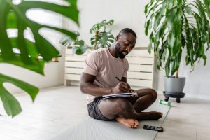 Young man sitting cross-legged on a yoga mat at home, writing in a notebook after a workout, with a smartphone nearby, creating a calm and focused atmosphere.