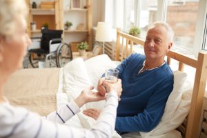 A senior man with gray hair is sitting up in bed, recovering from gallbaldder removal, and receiving a glass of water from another individual.
